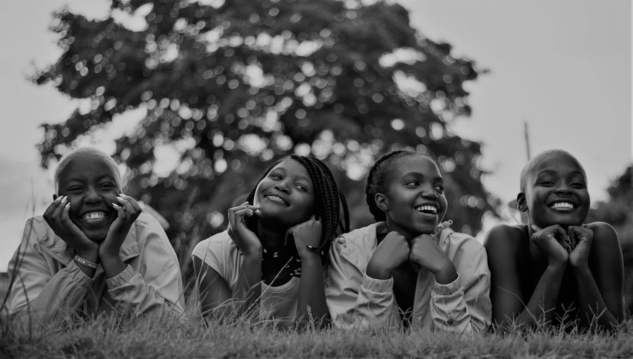 Contact A group of four happy black women lying on grass, enjoying a joyful moment outdoors.