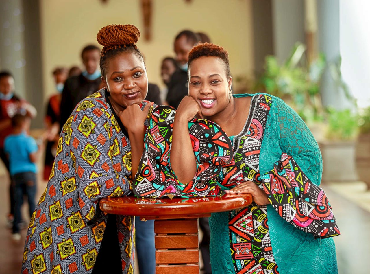 Two women posing in colorful African garments indoors, exuding joy and friendship.