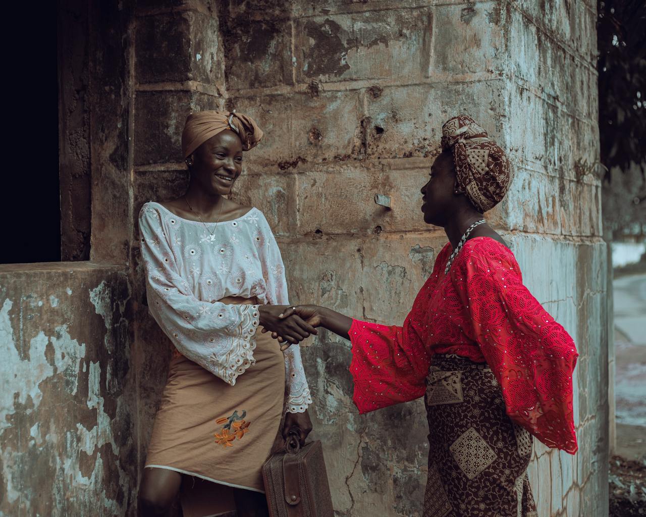 Two women in colorful traditional attire greet each other warmly by shaking hands.